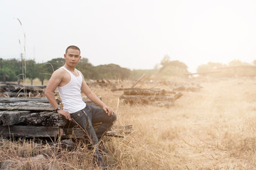 Naklejka na ściany i meble Asian young man in white vest and jeans sits on dry grass field