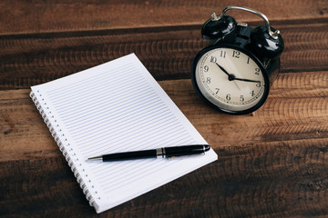 clock, pen and blank notebook on wooden table background