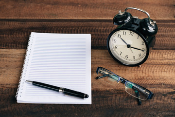 clock, glasses, pen and blank notebook on a wooden table background