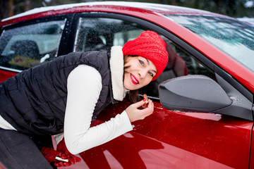 Girl in a red cap and warm jacket paints her lips near mirror of red car