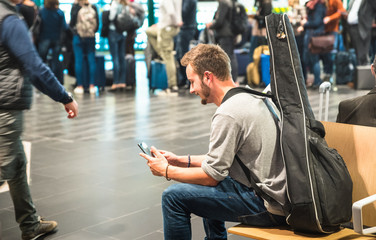 Hipster man at international airport using mobile smart phone - Wanderer person at terminal gate waiting for airplane - Wanderlust travel trip concept with guy and guitar backpack - Focus on face