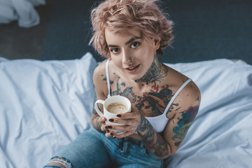 young tattooed girl with pink hair drinking coffee in bed at morning