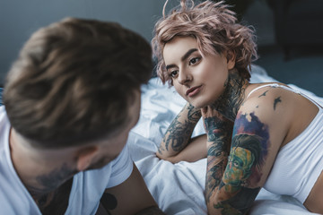 young tattooed couple looking at each other and lying on bed in bedroom