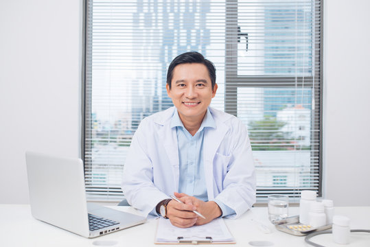 Smiling asian doctor sitting at his desk in medical office