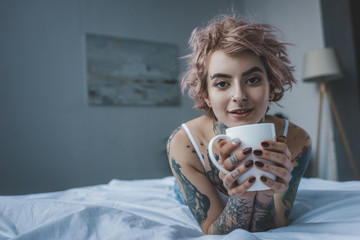 smiling tattooed girl with pink hair drinking coffee in bed at morning