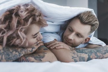 young tattooed couple looking at each other under white blanket in bedroom