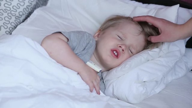Sick child boy laying in bed and mom hand taking temperature. Mother checking temperature of her sick son. Sick child with fever and illness in bed at home.
