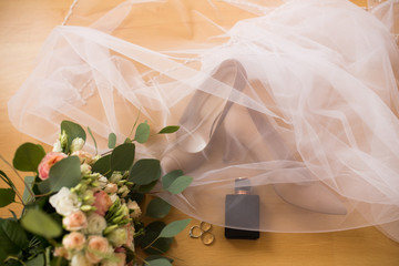 Wedding accessories. Black perfume, wedding rings, beige bride’s shoes are covered the veil on a wooden background. 