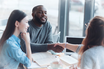 Good job. Confident international male person leaning elbows on the table while holding hands with colleague and keeping smile on his face