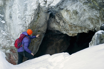 girl hiker in winter peeks into a dark cave in a steep limestone rock
