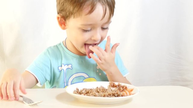 A child is eating buckwheat porridge with meat, sitting at a table on a white background.