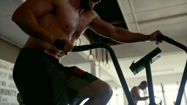 Muscular athlete doing intense workout on gym bike. Fitness man spinning stationary bicycle in cross fit gym.