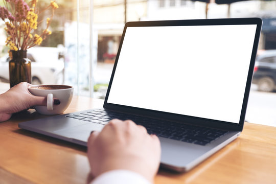 Mockup image of a businesswoman's hand using laptop with blank white desktop screen while drinking hot coffee on wooden table in cafe