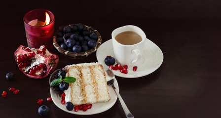 Still life Piece of layer cake with blueberries, chocolate cookies and pomegranate. Dark wooden background. Burning candle. Romantic Valentine's Day concept. Selective focus.