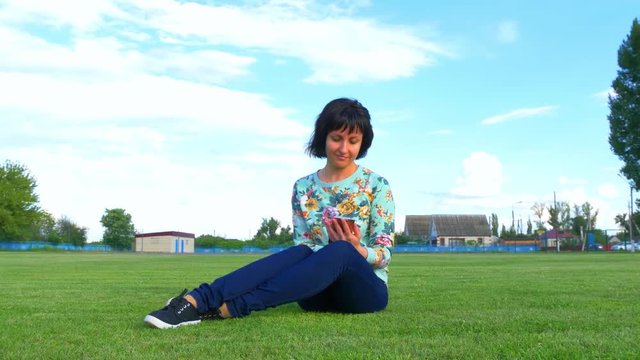 Young woman sitting on the lawn of the stadium uses a smartphone on a cloudy blue sky background.