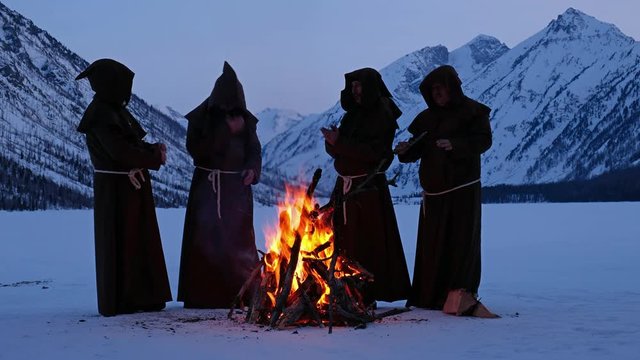 Group of monks in brown cloaks of the order of St. Francis are warming themselves by the fire