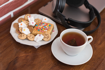 Sweets and cookies with cheerful faces, a cup of fragrant tea behind a laptop and wireless headphones on a mahogany table