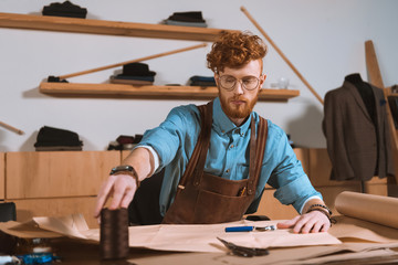 young male fashion designer in apron and eyeglasses sitting at workplace