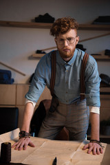 handsome young fashion designer in eyeglasses looking at camera while leaning at table with craft paper