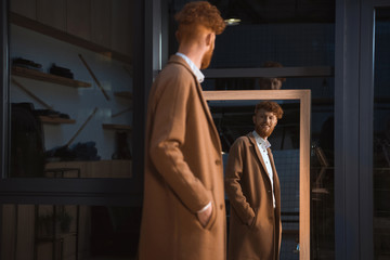 smiling young man wearing overcoat and looking at mirror in boutique