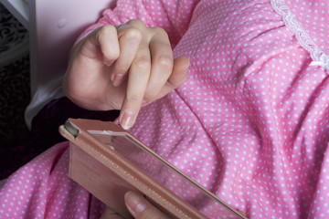 A woman in a nightgown works with a tablet while lying in bed. She touches the screen with her fingers.
