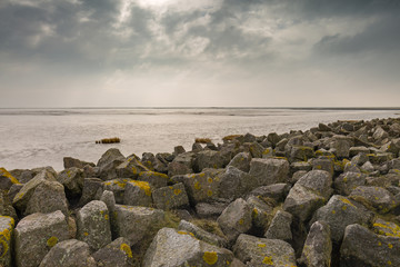 Fototapeta na wymiar The clouds convey a threatening mood over the calm North Sea at low tide in the Watt. Artificially piled rocks protect the coast from storm surges - Mehldof Bay, Mehldorf, Germany