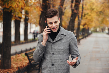 Picture of elegant male in coat walking in empty park with autumn trees, and talking on smartphone