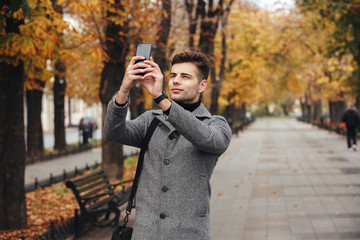 Handsome man in coat taking photo of beautiful autumn trees using his modern cellphone while walking in empty park