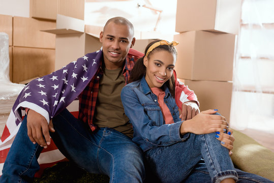 african american couple with united states flag sitting on floor at new home with cardboard boxes