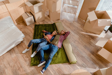 top view of dreamy african american couple lying on floor in new home with cardboard boxes