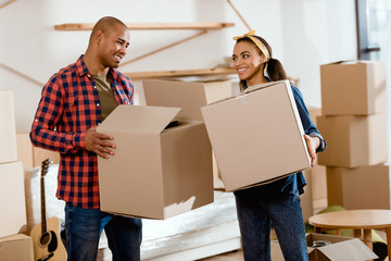 smiling african american couple holding cardboard boxes and moving to new home