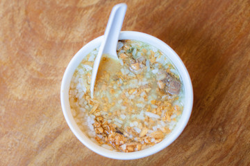 Boiled rice Asain Thailand style breakfast food in the morning, boiled rice wiht pork and deep fried garlic in the white bowl and white spoon put on the wood table
