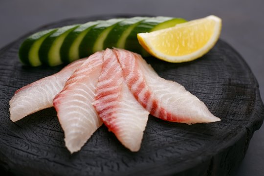 Red sea bream sashimi with cucumber and lemon slices on dark wooden background, close up. Traditional Japanese food