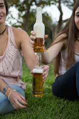 Two young laughing women on lawn in park toasting with beer bottles having fun. 