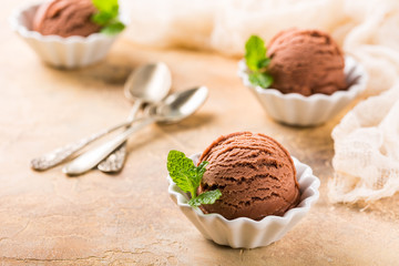 Chocolate ice cream scoop in white bowl with mint leaves on sand color stone background. Summer food concept with copy space.