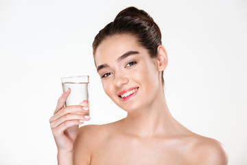 Close up picture of glad gorgeous woman being half-naked drinking minaral water from transparent glass with smile over white background
