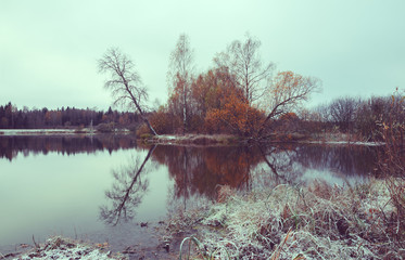 Naklejka na ściany i meble Cloudy autumn landscape with trees growing on the bank of pond.First snow.Overcast sky.Vintage colors.