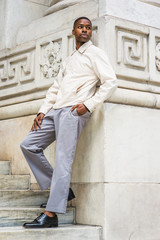Obraz premium Portrait of Young Handsome African American Man in New York. Young black man wearing light color jacket, gray pants, black leather shoes, standing against vintage marble wall, looking around, relaxing