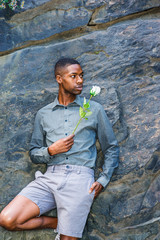 Naklejka premium Young African American Man missing you and waiting for you. Young black man wearing gray shirt, shorts, standing against rocky wall at Central Park in New York, holding white rose, looking around..