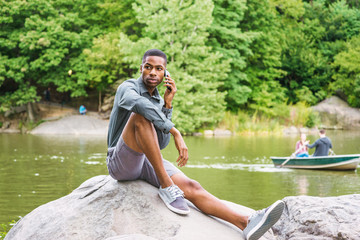Young African American Man wearing gray long sleeve shirt, shorts, sneakers, sitting on rocks by lake at Central Park in New York, relaxing, talking on cell phone. People rowing boats on background..