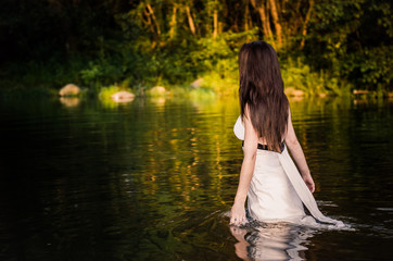 A long-haired fit girl with white dress walking through the river touching water