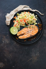Grilled salmon and cuscus with vegetables served on a metal tray. Flat-lay on a dark brown stone background, vertical shot with copy space