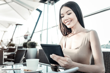 Helpful gadget. Nice-looking stylish girl sitting by the table holding the tablet and looking at screen.