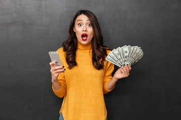 Shocked brunette woman in sweater holding money and smartphone