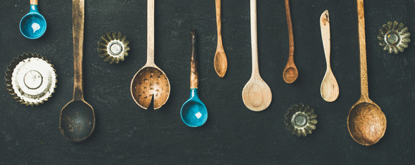 Flat-lay of various old vintage kitchen spoons and baking tin molds over black stone background, top view, copy space, wide composition. Rustic cooking concept