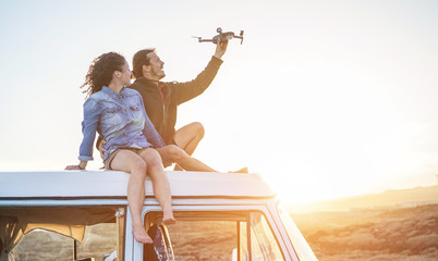 Happy couple using a drone sitting on top of vintage minivan at sunset - obrazy, fototapety, plakaty