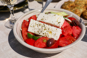 A bowl of village greek salad next to zucchini balls plate and white wine, water glasses served in greek tavern.