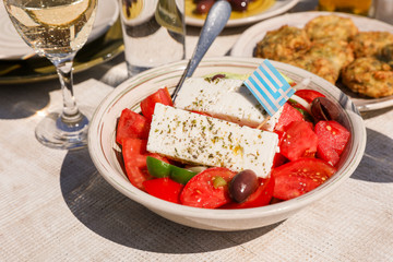 A bowl of village greek salad with greek flag on next to zucchini balls plate and white wine, water glasses served in greek tavern.