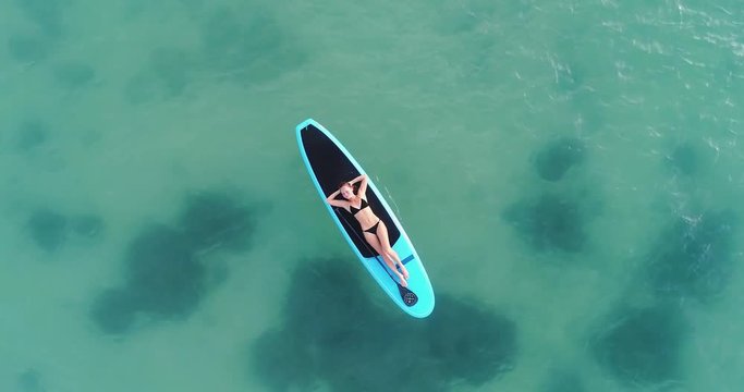 Camera moving around Attractive woman in bikini is sunbathing on a surfboard, aerial view