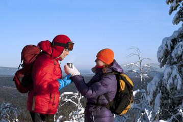 Two hikers on top of the mountain on a clear frosty day against the backdrop of a forest landscape in the valley; a woman offers a man a hot tea in a cup (in a thermos cap).